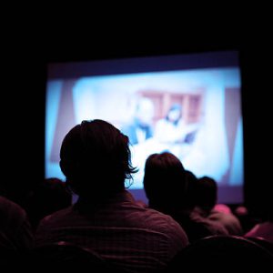 18 filmabende in deutsch und spanisch an verschiedenen Terminen (ES und DE) An unknown number of people sitting in a dark theater. There is a projector screen in front of them with a man and woman at a conference.