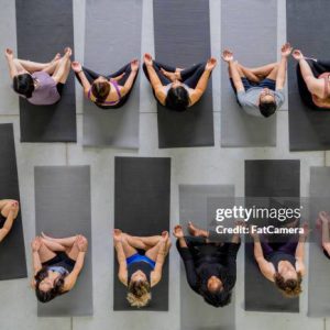 5 yoga übers internet (ES und DE) A large group of adults are seen each sitting out on a yoga mat in this aerial view. They are all wearing fitness attire and are holding a lotus pose as they meditate.